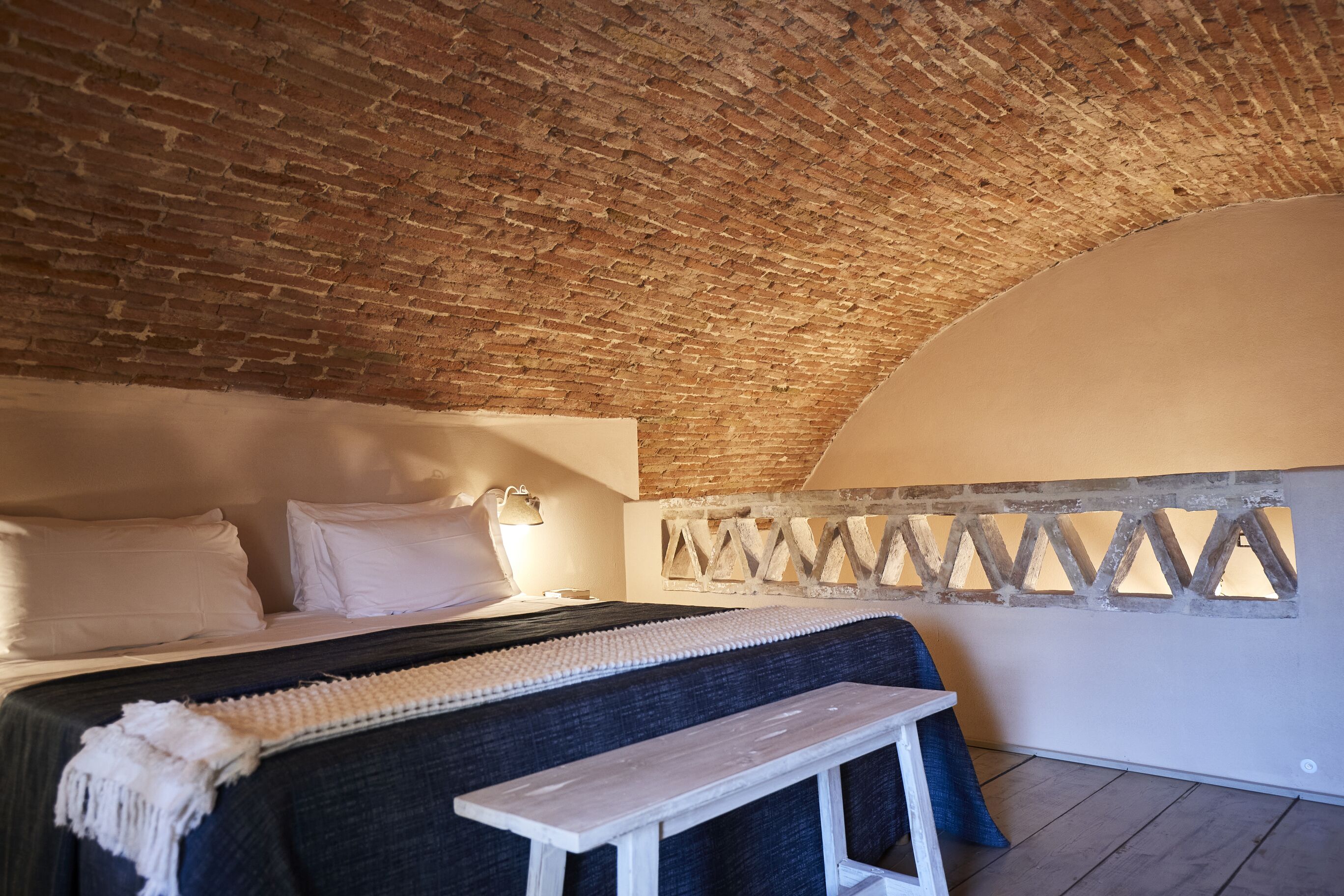 Un dormitorio acogedor con techo de ladrillo, cama con ropa blanca, una banca y una lámpara de pared.
