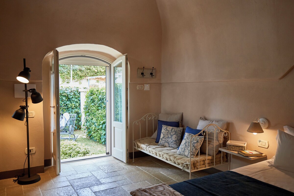 A cozy bedroom with a bed, bedside table, lamp, and a view of the garden through an open door.