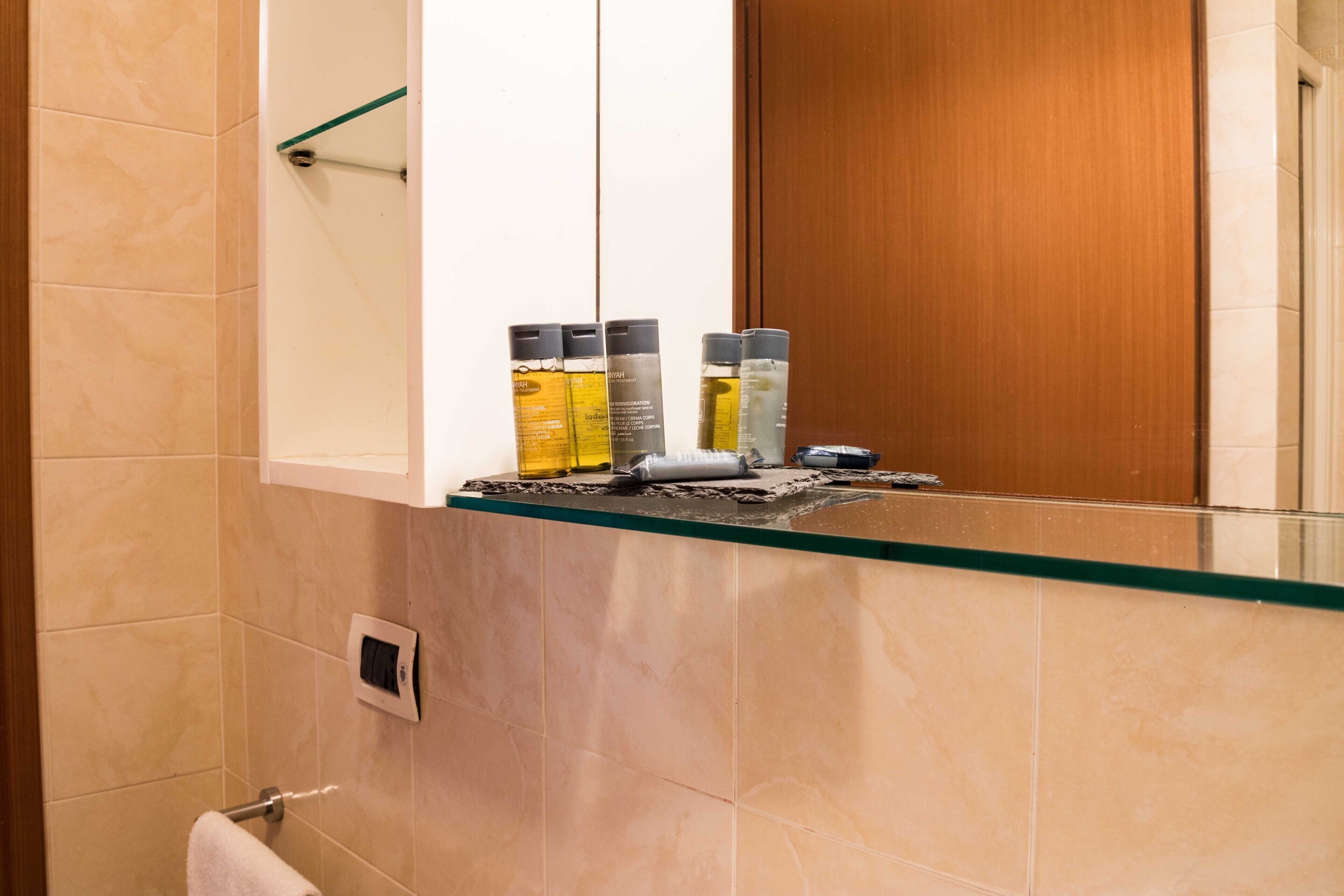 Une salle de bain carrelée de carreaux beiges, une étagère en verre où sont rangés les produits de toilette, et une porte en bois.
