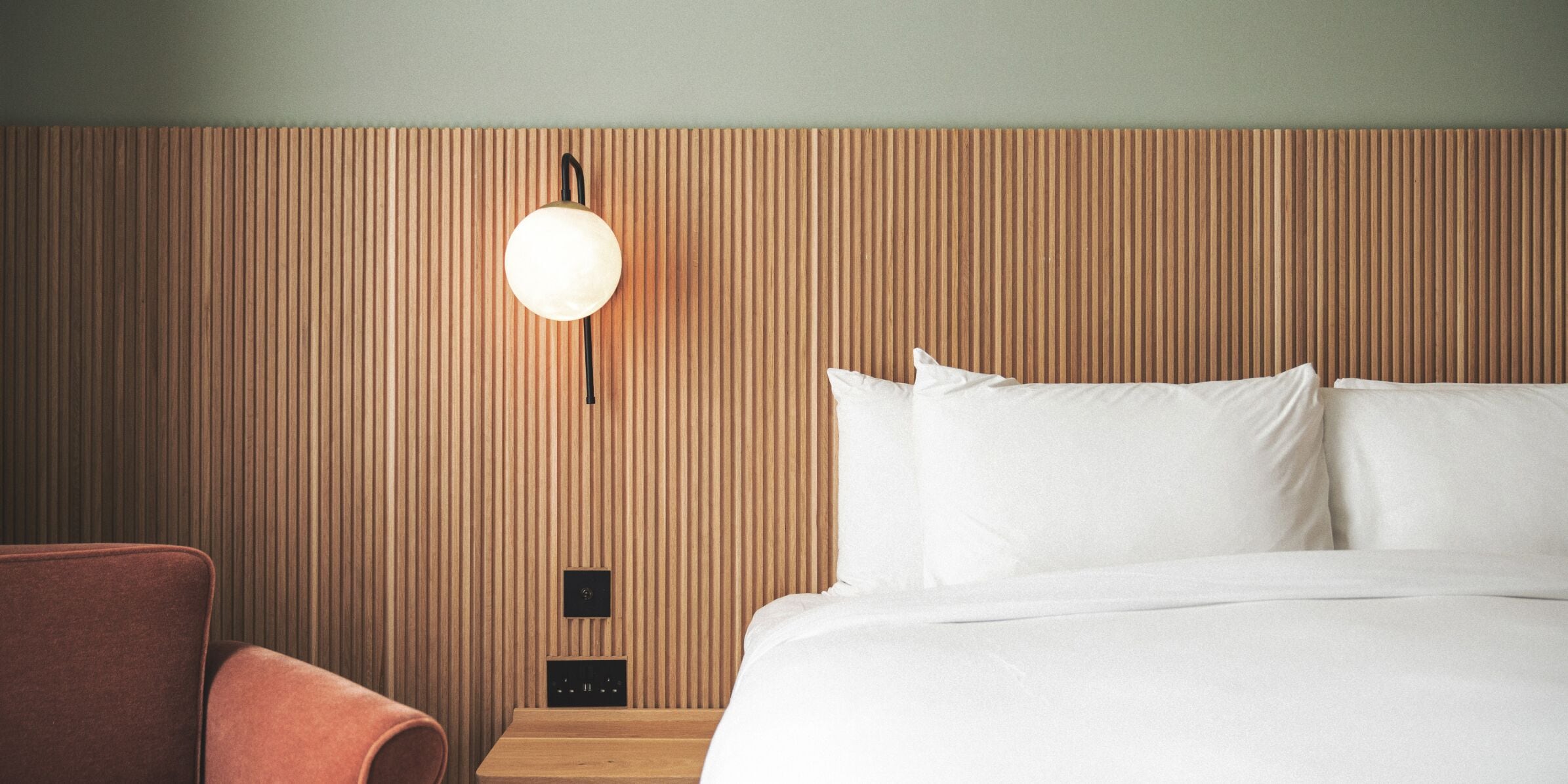A hotel room with a wooden headboard, a bed with white linens, a round wall-mounted light, and a small wooden nightstand.