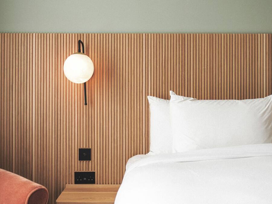 A hotel room with a wooden headboard, a bed with white linens, a round wall-mounted light, and a small wooden nightstand.