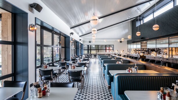 L’intérieur d’un restaurant se caractérise par de hauts plafonds, de grandes fenêtres et un sol à motifs. Des tables et des banquettes sont disposées pour accueillir les clients.