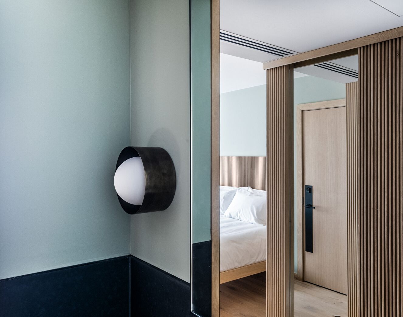 A hotel room with a bed, a wall-mounted light, and a wooden wardrobe.