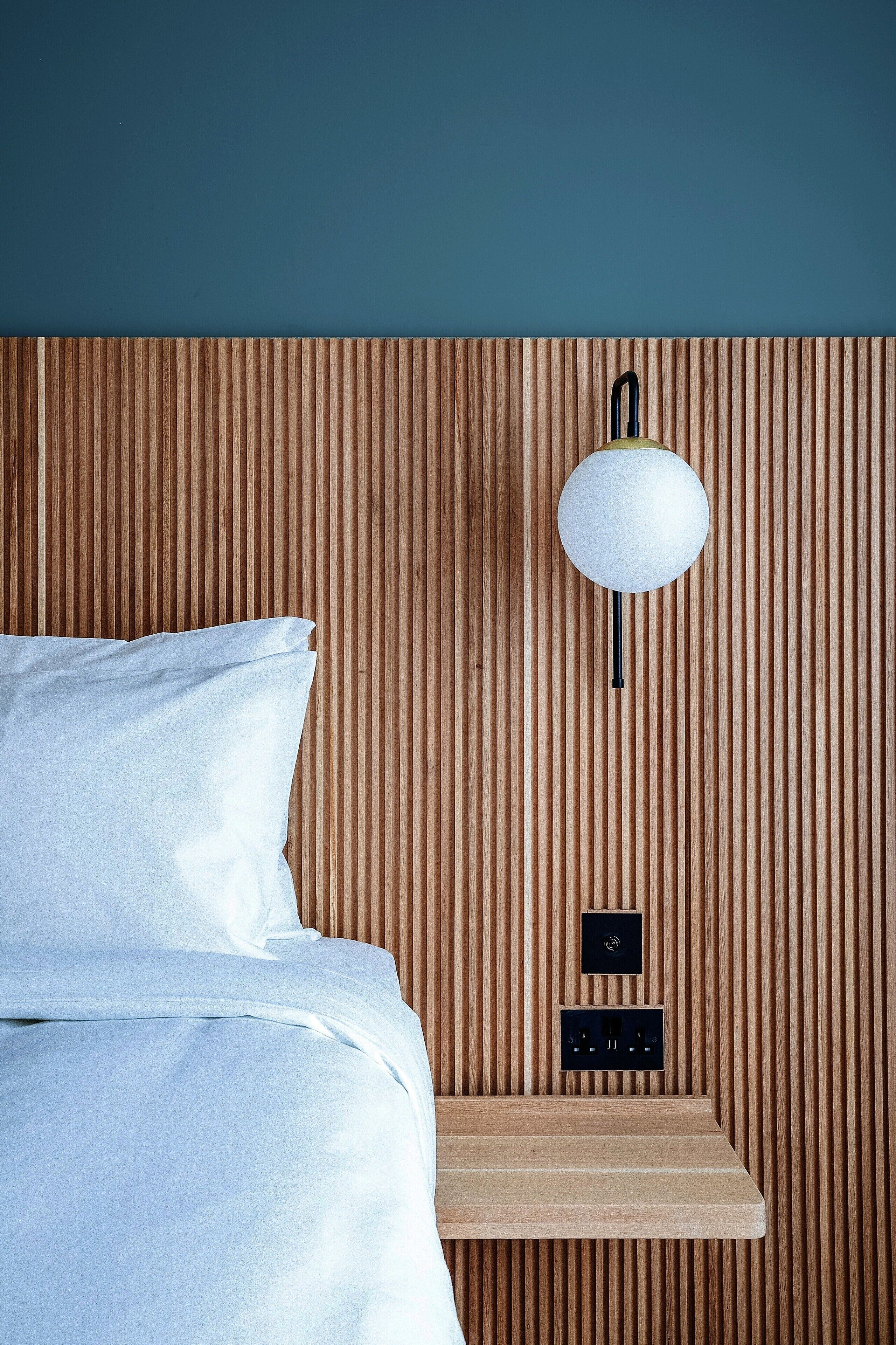A bed with white bedding, a wooden slatted headboard, a wall-mounted light fixture, and a small wooden shelf with electrical outlets.