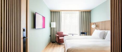 A hotel room with a bed, a chair, a small table, a lamp, and a painting on the wall.