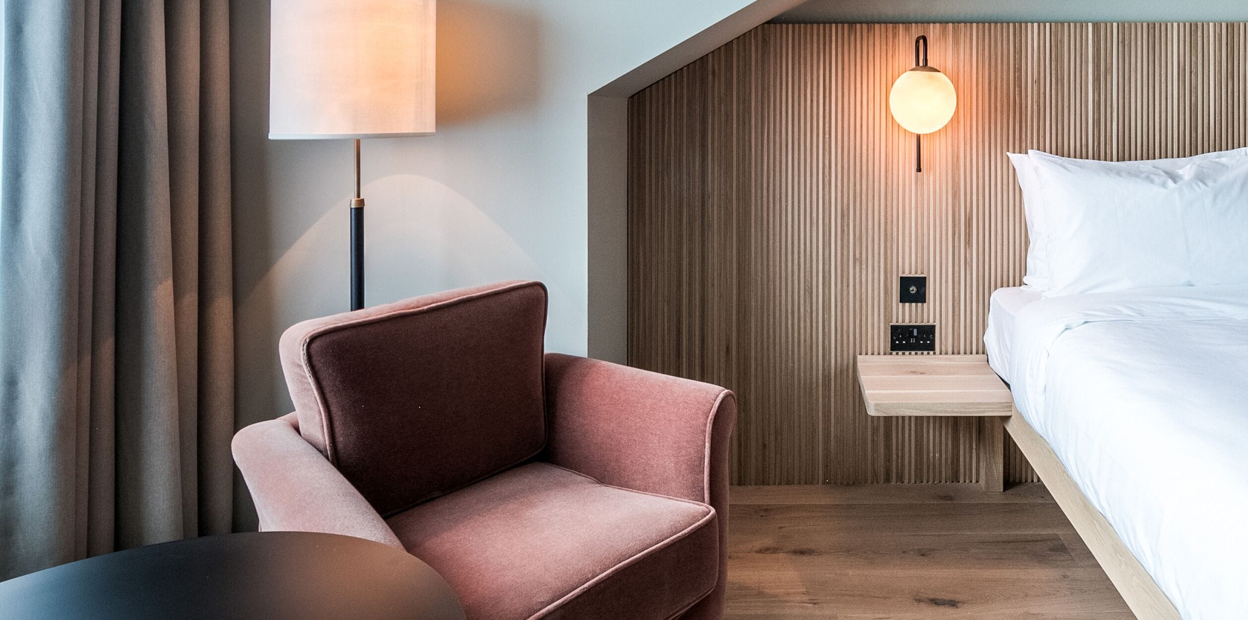 A modern hotel room with a wooden wall panel, a bed with white linens, a pink armchair, a bedside table with a lamp, and a power outlet.