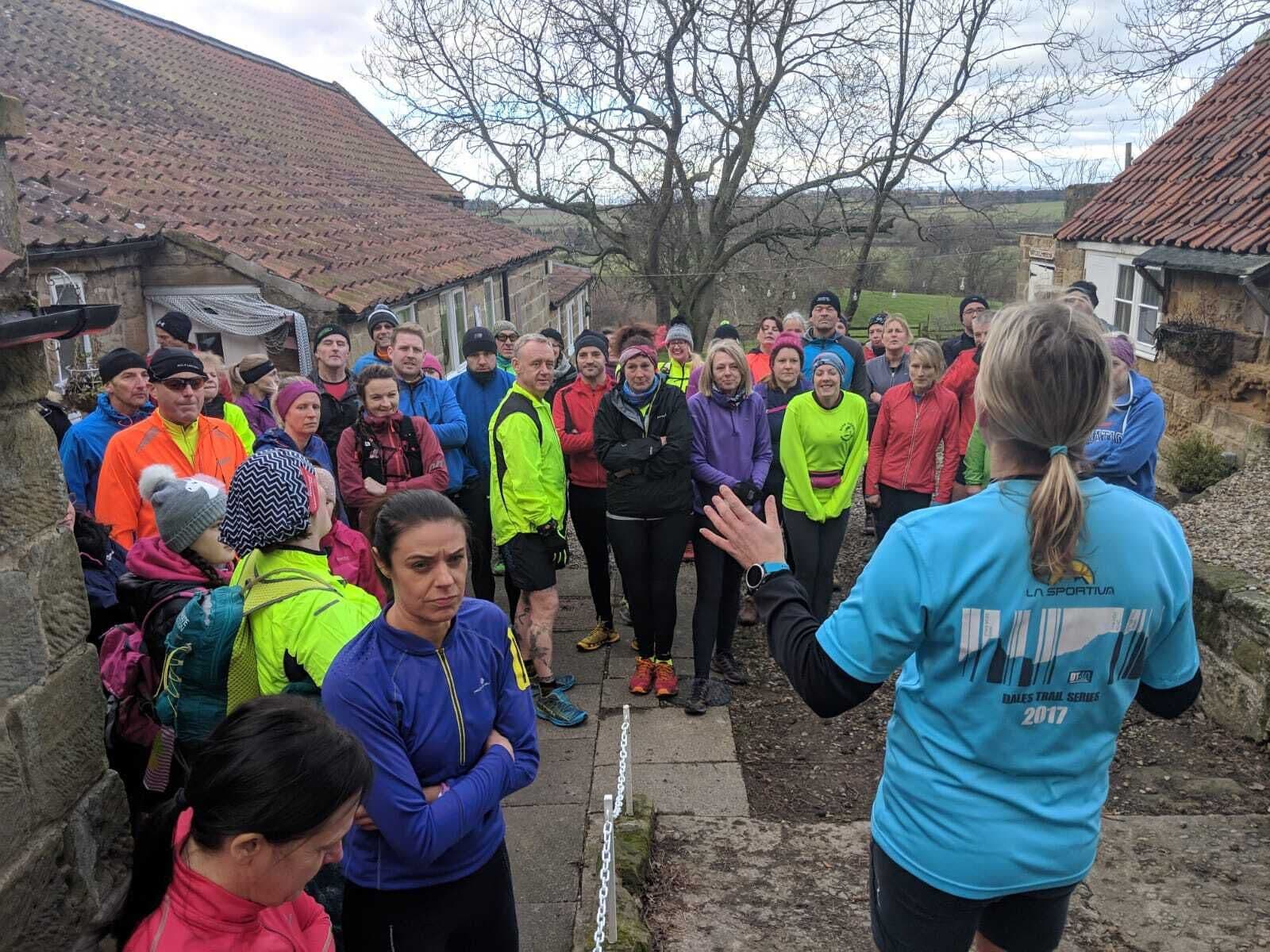 Un grupo de personas reunidas al aire libre, posiblemente en un evento comunitario o una reunión, con edificios de piedra y un árbol al fondo.