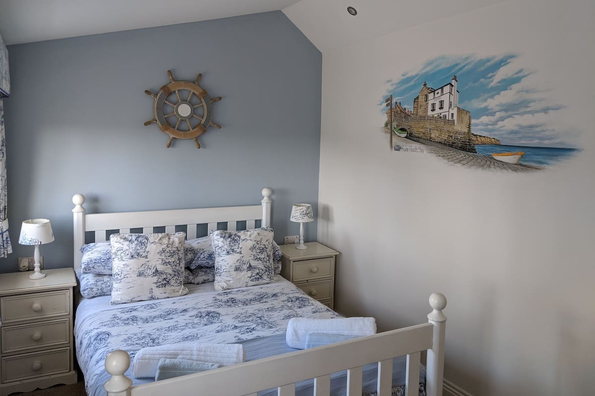 Un dormitorio con cama, mesitas de noche, una rueda de barco montada en la pared y un mural con escena costera.