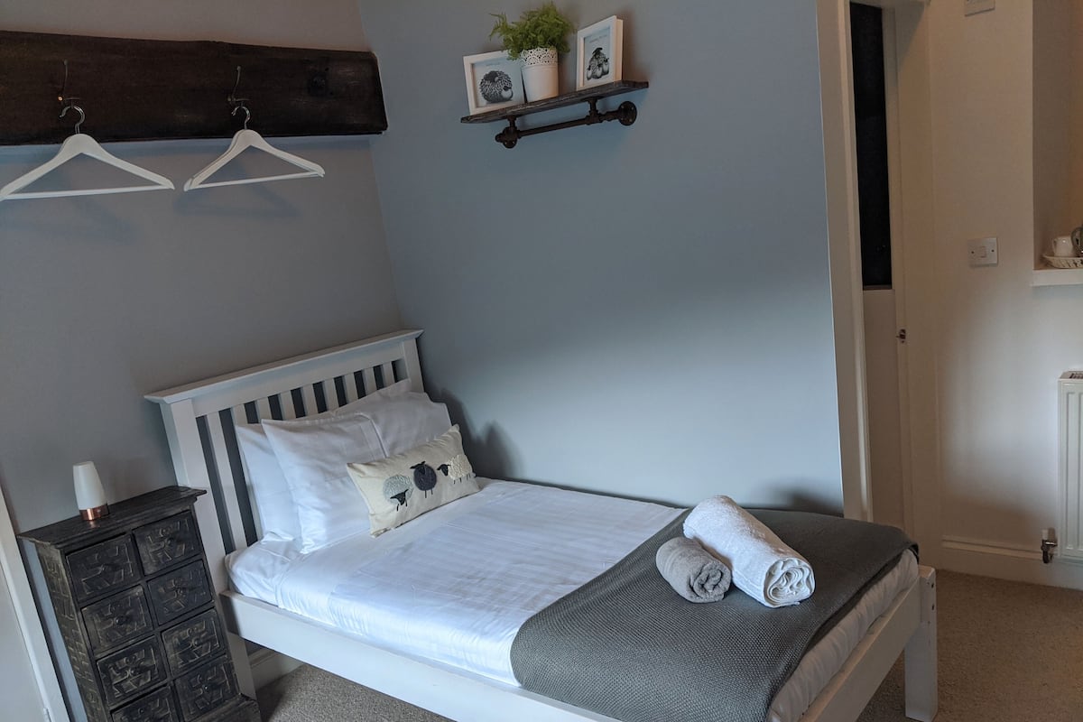 Una cama bien hecha con almohadas y una colcha, un aparador con una lámpara, y un estante con objetos decorativos.