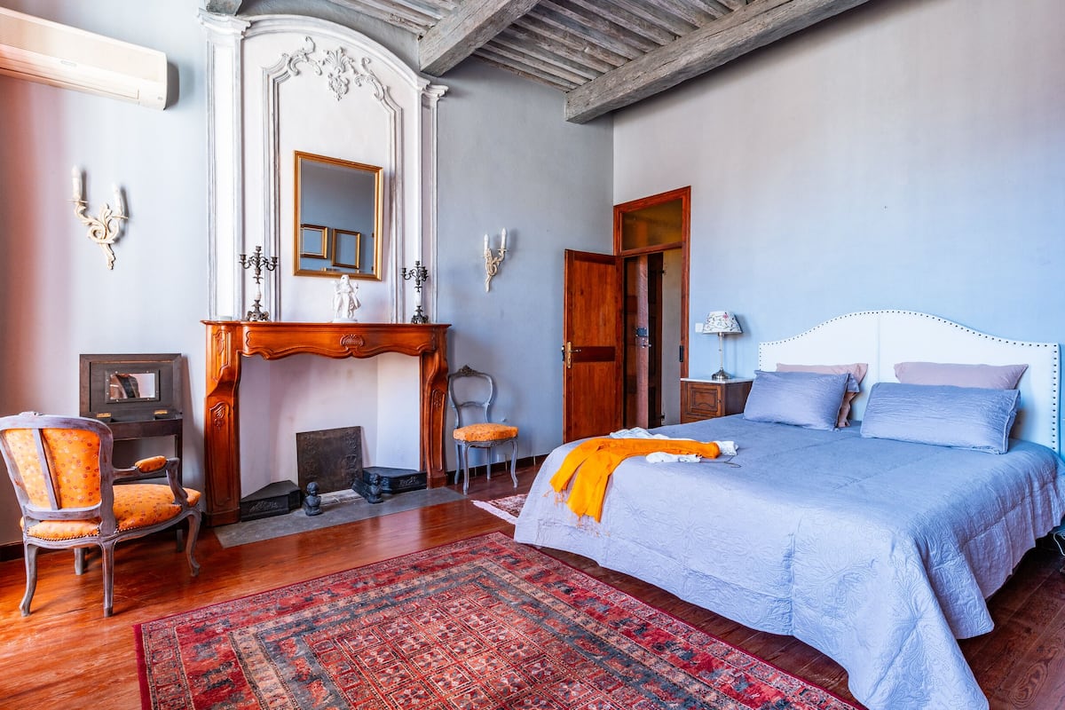 Une chambre à coucher avec un grand lit, une commode en bois, une chaise, une cheminée et un miroir.