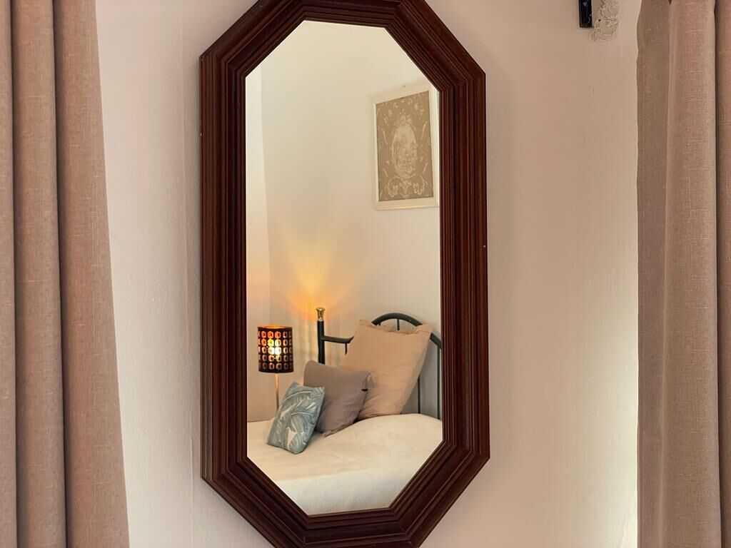 Un dormitorio con un espejo octogonal grande, una cama con almohadas, una mesita de noche con lámpara y un cuadro enmarcado en la pared.