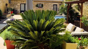 Une cour agrémentée d’un grand palmier, d’un espace aménagé pour s’asseoir à l’extérieur et d’un bâtiment en pierre surmonté d’un toit en tuiles.
