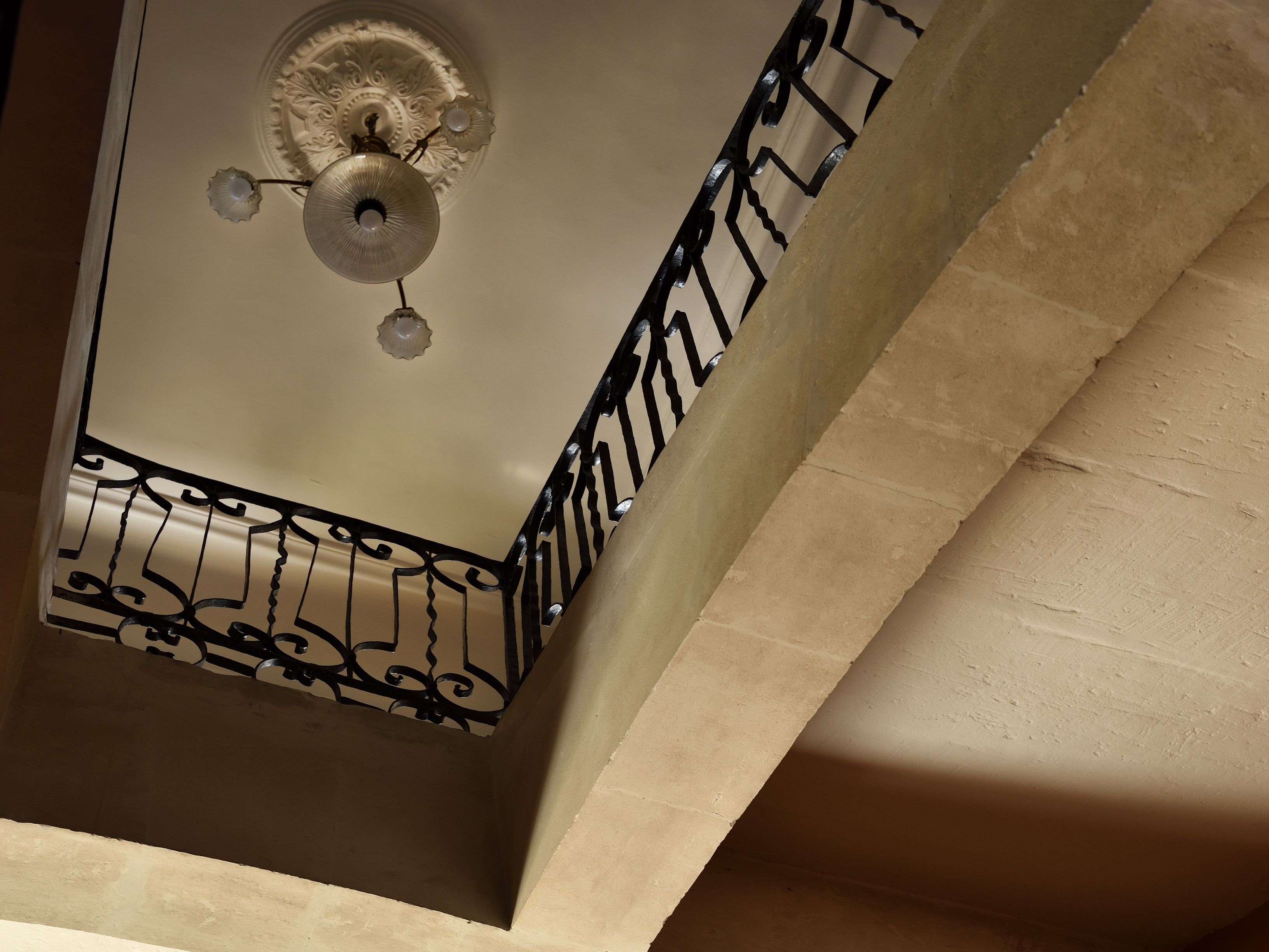 Een hoek van een kamer met een decoratief plafond, een zwarre metalen balustrade en een lamp.