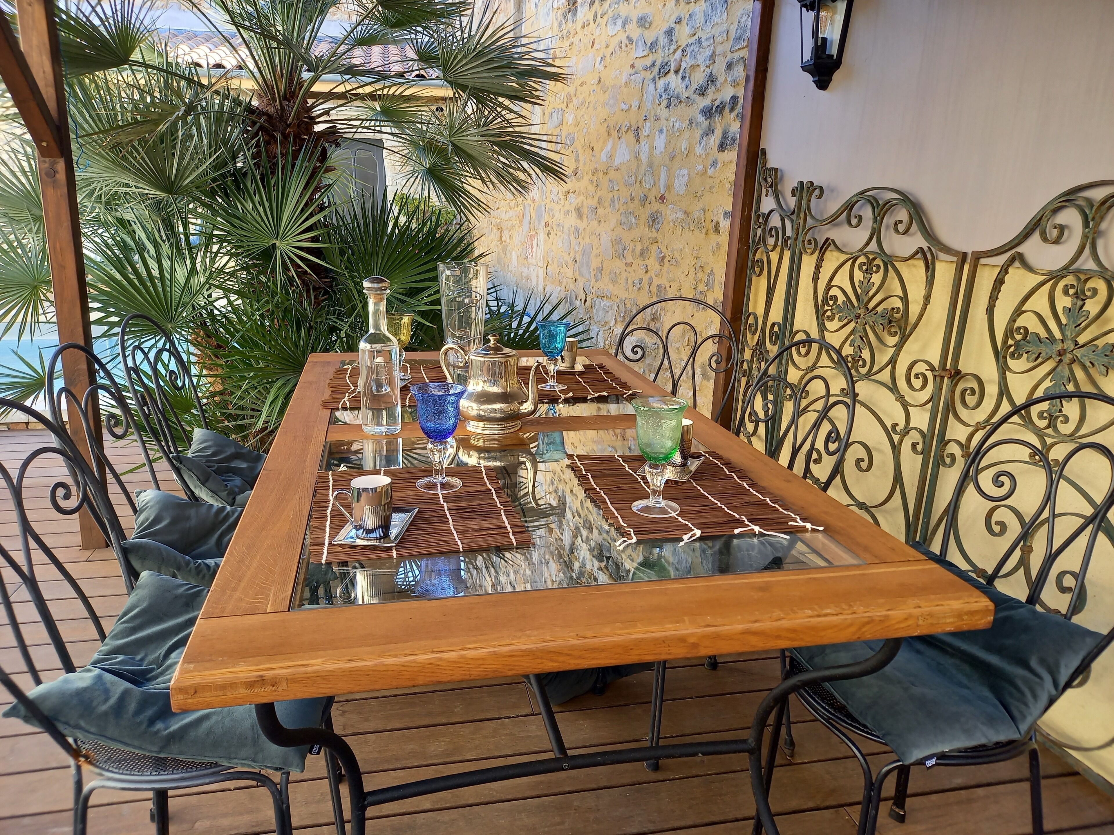 Una mesa con cubierta de vidrio y sillas de metal, frente a una pared de piedra con baranda de hierro forjado.