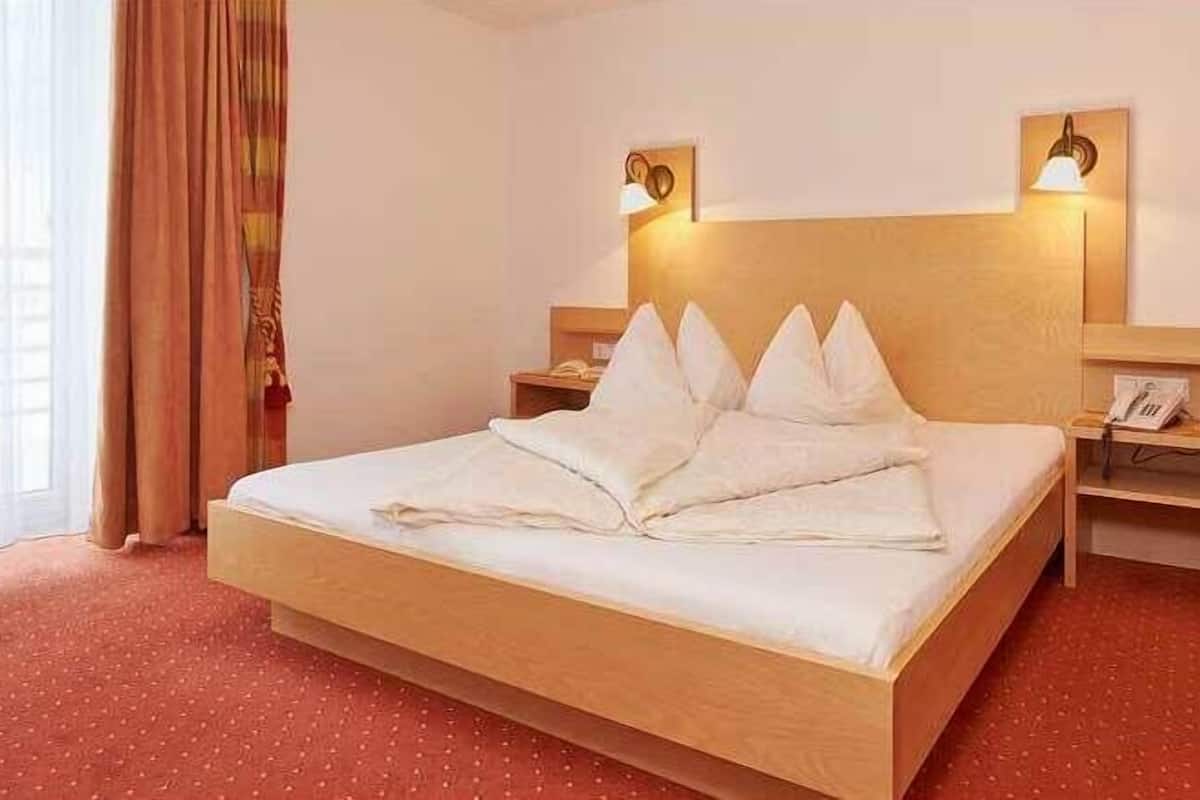 A hotel room with a large bed, two bedside tables, a nightstand, a window with curtains, and a wall-mounted lamp.