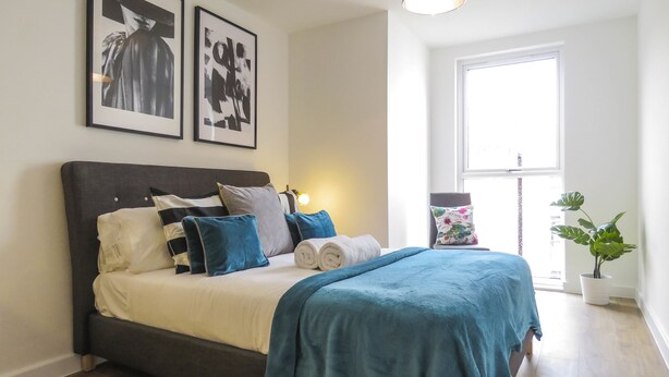 A neatly made bed with a dark headboard, multiple pillows, and a blue blanket. There are two framed pictures on the wall above the bed. - Tudors eSuites Birmingham Canalside Apartments (Birmingham)