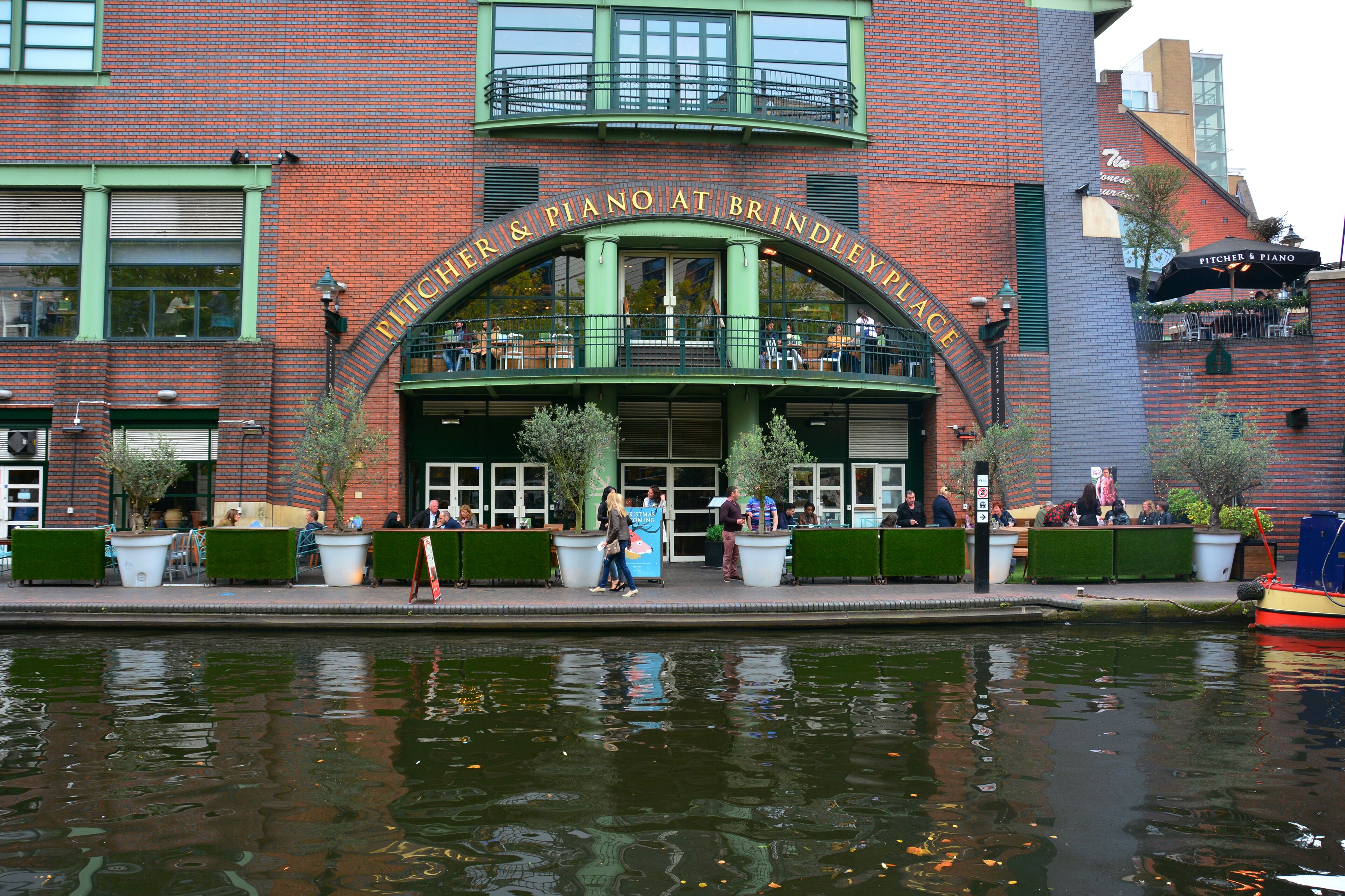 Un bâtiment en briques avec une arche verte portant l’inscription « PITCHER & PIANO AT BRINDLEY PLACE » et des tables à l’extérieur, agrémentées de plantes en pot.