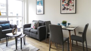 A modern living room with a grey sofa, a small round table, and a dining table with chairs. There are two framed artworks on the wall and a window providing natural light. - Tudors eSuites Birmingham Canalside Apartments (Birmingham)