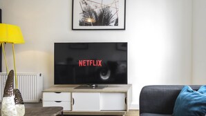 Un salon moderne avec un téléviseur à écran plat affichant le logo de Netflix, un meuble en bois en dessous, une lampe de sol jaune et un coussin bleu sur un canapé foncé.