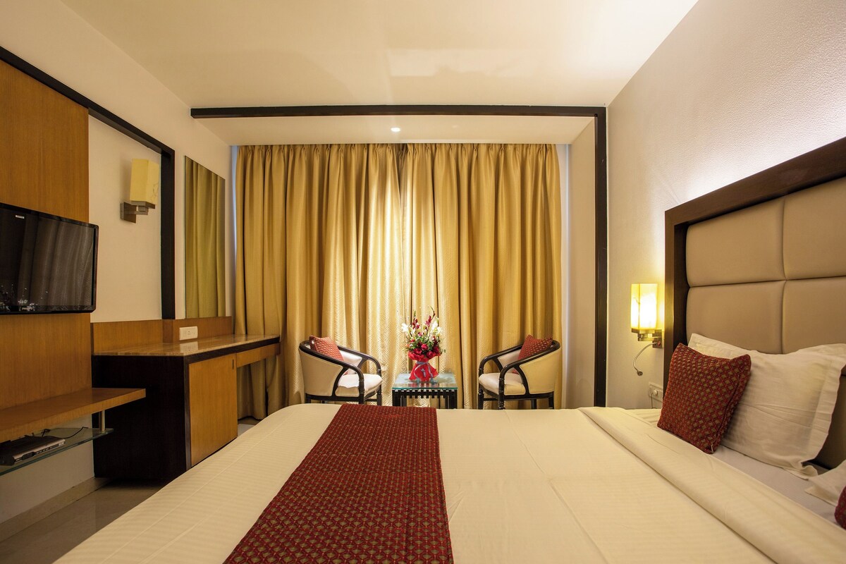 A hotel room with a bed, a desk, two chairs, a TV, and a window with curtains.