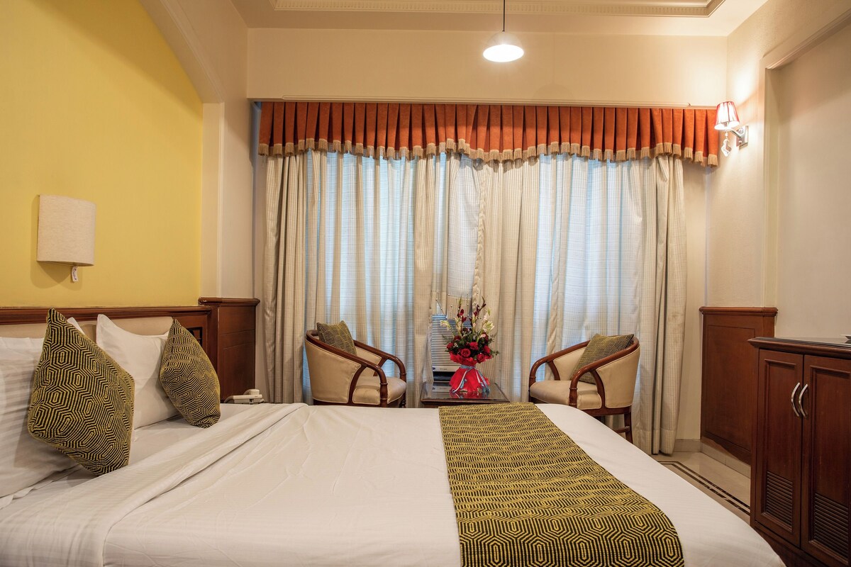 A hotel room with a bed, two armchairs, a nightstand with a vase of flowers, a lamp, and a window with curtains.