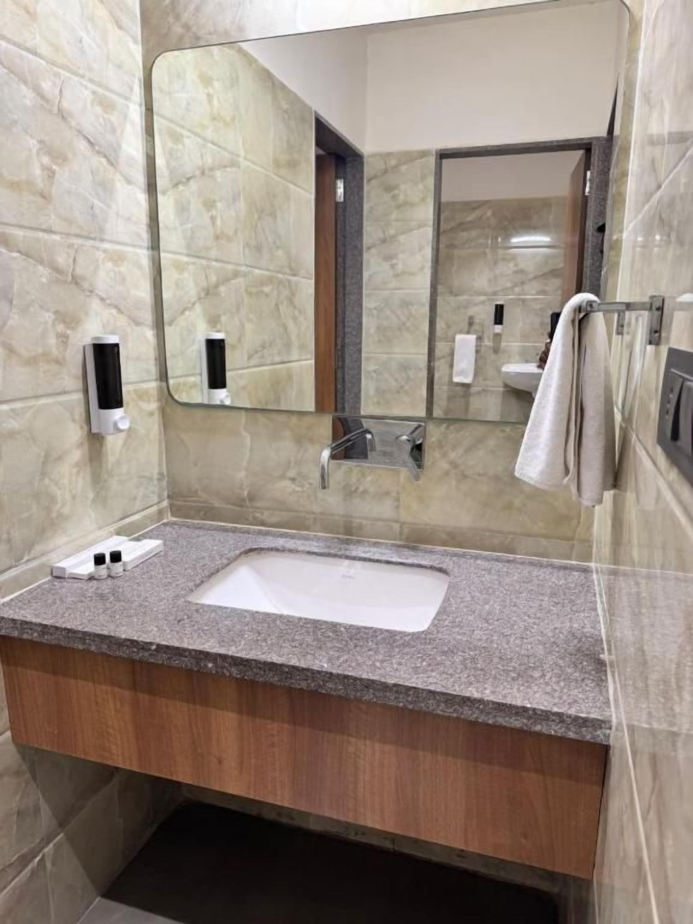 A bathroom with a granite countertop sink, a mirror, a towel rack, and a soap dispenser.