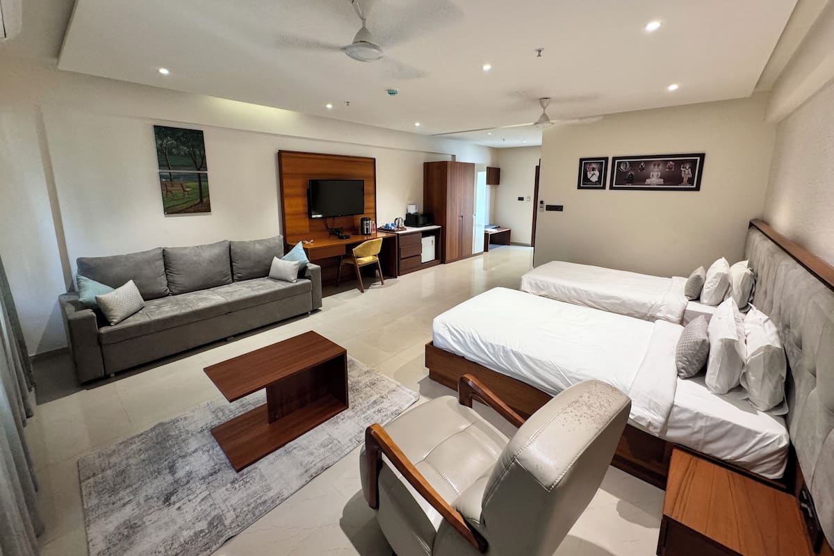 A modern hotel room with a sofa, two single beds, a desk, a chair, and a television.
