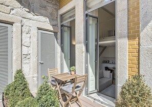 Un patio al aire libre con una mesa y sillas de madera, una planta en maceta y vistas al interior de una habitación con cocina y sofá.