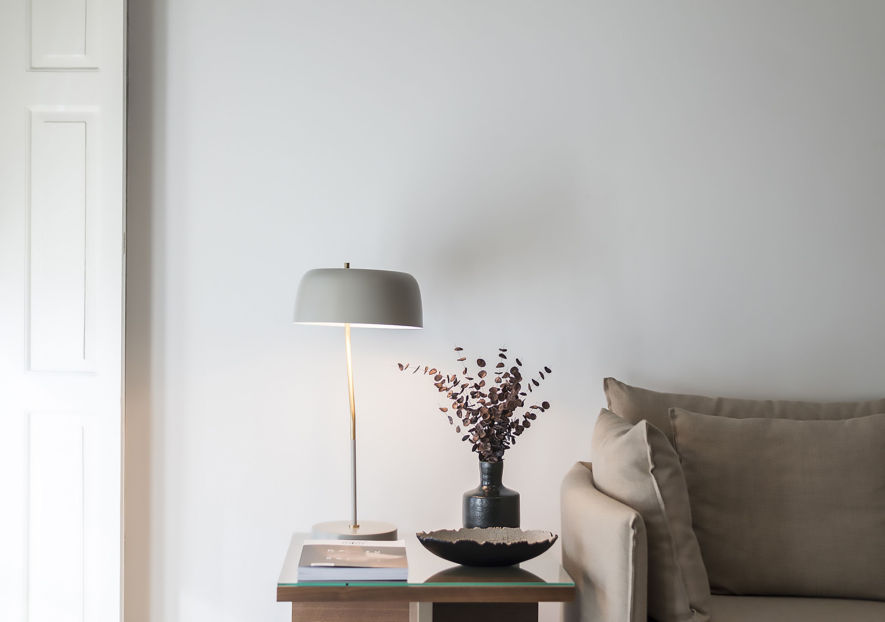Una sala de estar moderna con un diseño minimalista, que incluye un sofá, una mesita de noche con una lámpara y un jarrón con flores secas.