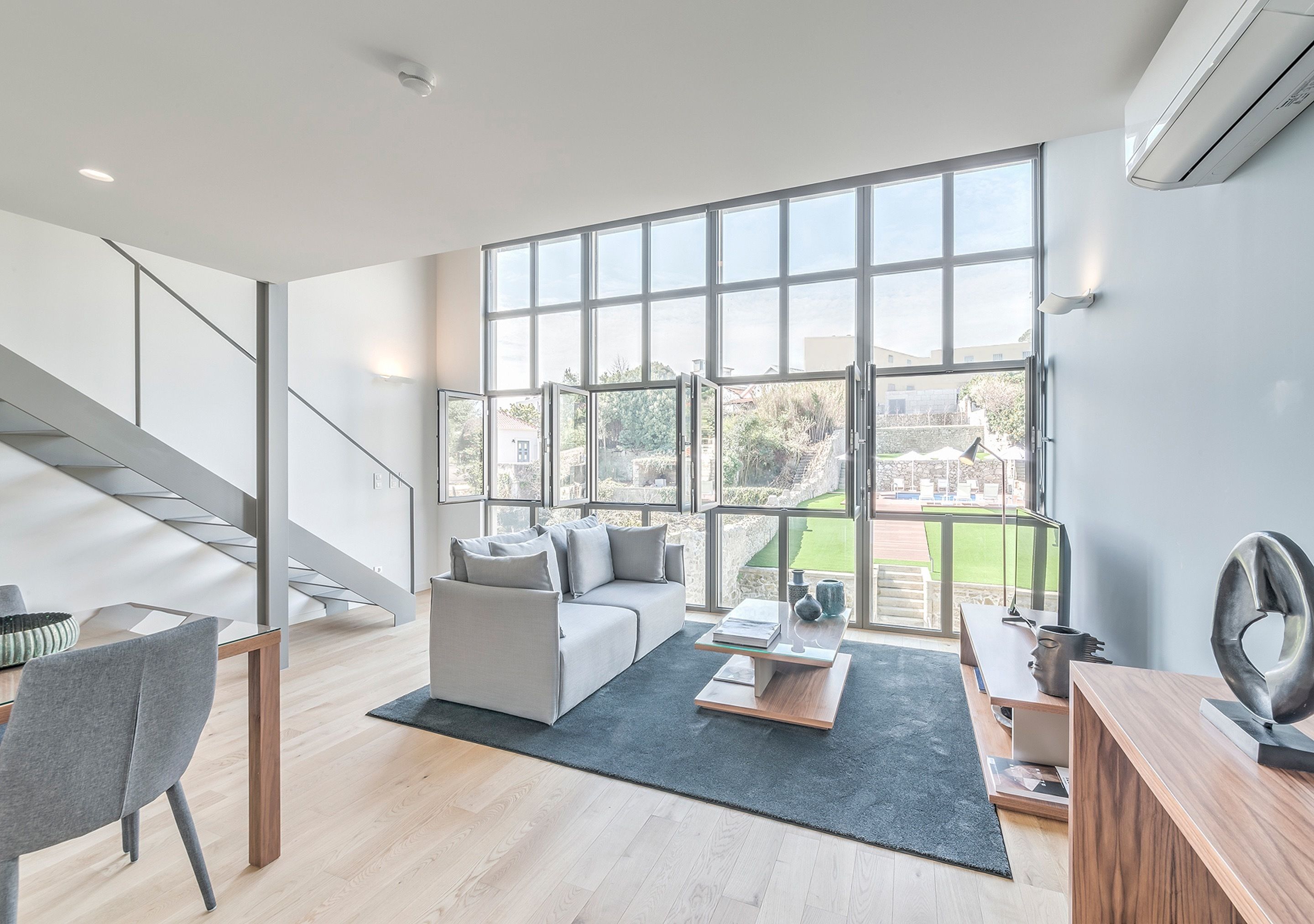 A modern living room with a large window, a grey sofa, a coffee table, and a staircase.
