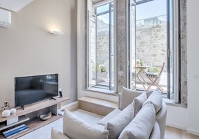 Una sala de estar moderna con un televisor de pantalla plana, un sofá y un área de comedor visible a través de ventanas amplias.