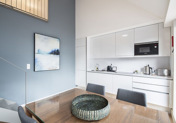 Una cocina moderna con armarios blancos, un microondas y una cafetera. Hay una mesa de comedor con sillas y una superficie de vidrio.