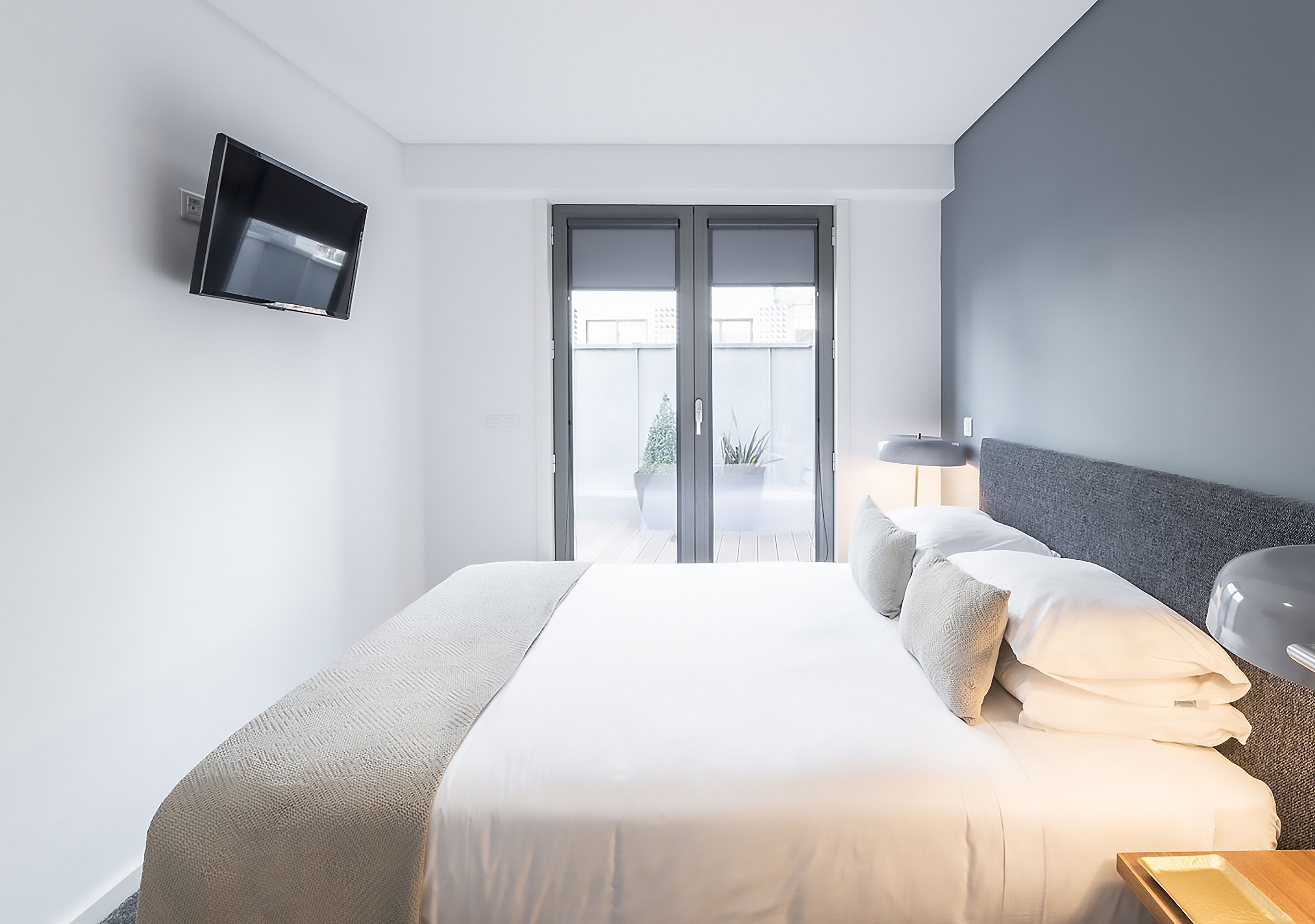 A modern hotel room with a large bed, a flat-screen TV mounted on the wall, a bedside table, and a view of the outdoors through a sliding door.