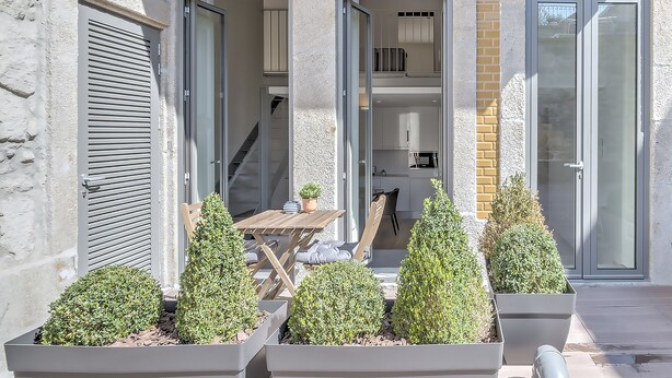 A modern building with a patio area featuring a table and chairs, surrounded by potted plants.