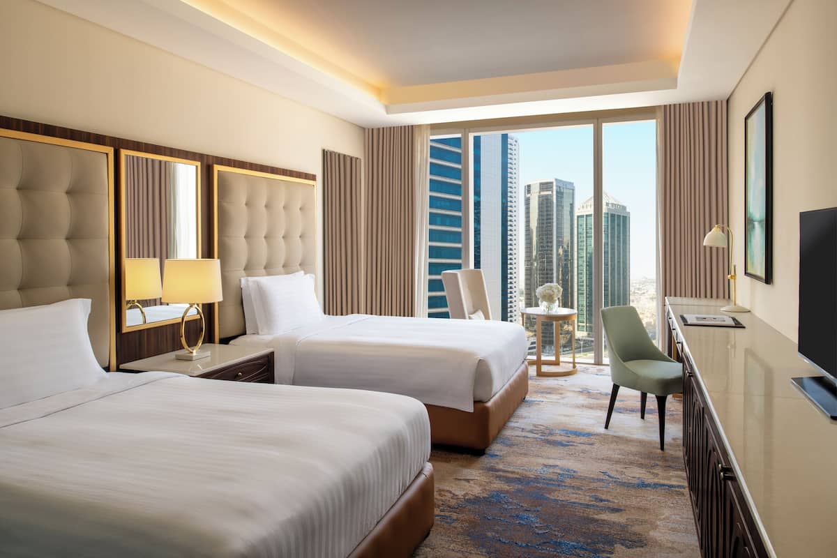 A hotel room with two beds, a desk with a TV, a chair, and a view of tall buildings.