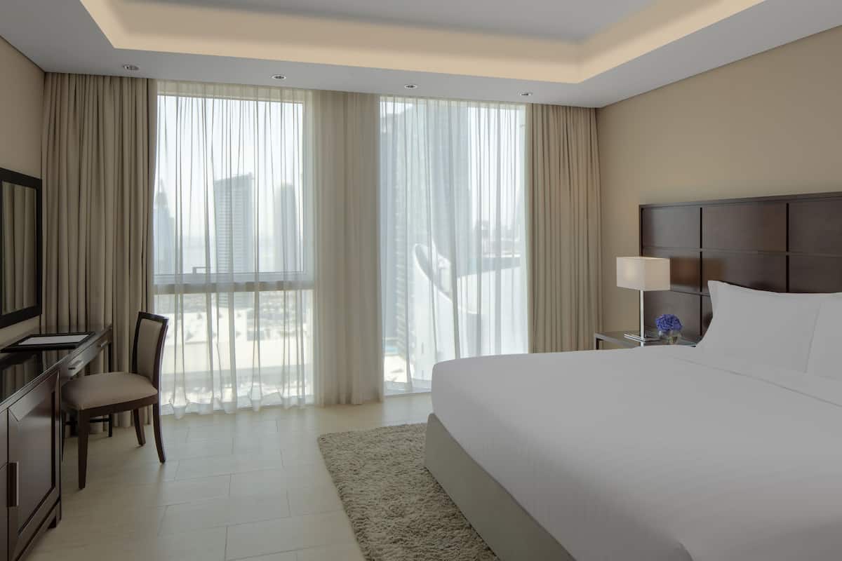 A hotel room with a large bed, a desk with a chair, a TV, and a window with sheer curtains.