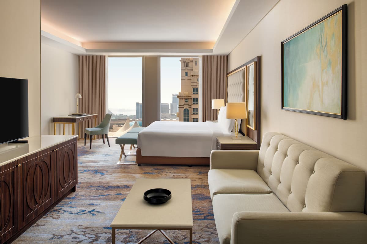 A hotel room with a large bed, a sofa, a desk with a TV, a chair, and a view of the cityscape.
