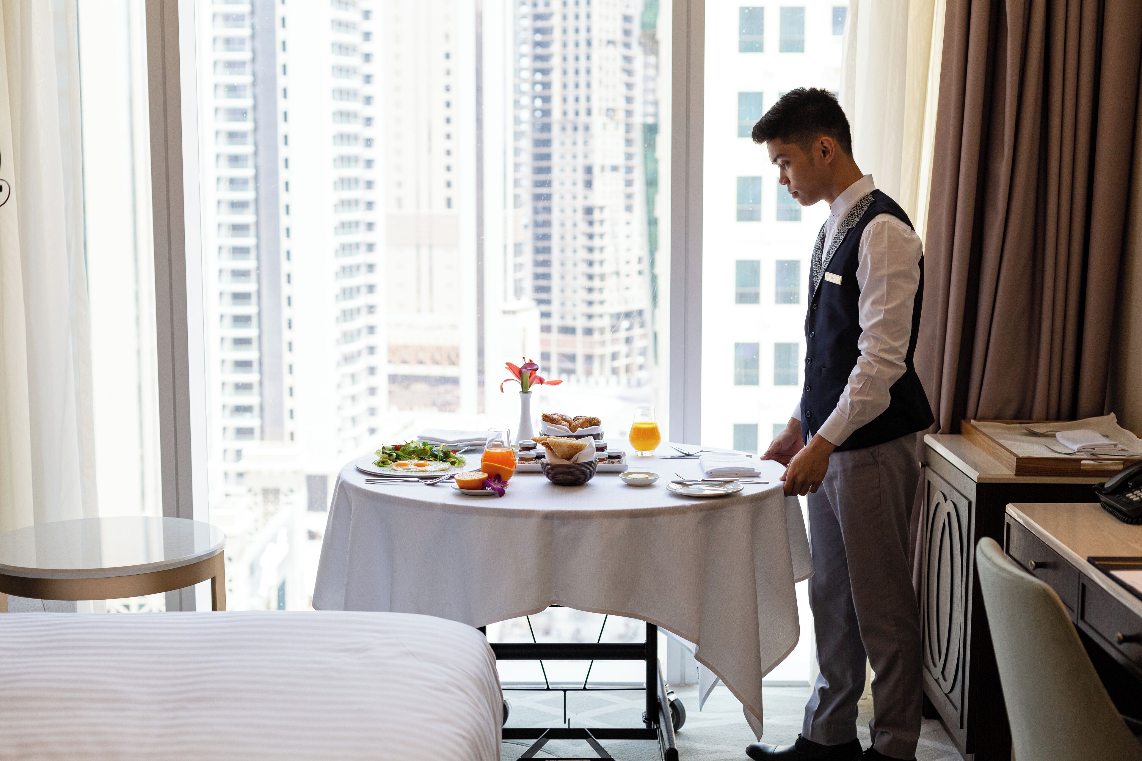 A hotel room with a dining table set for breakfast, a bed, and a view of tall buildings.