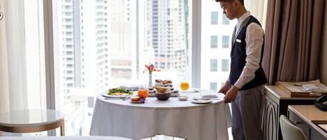 A hotel room with a dining table set for breakfast, a bed, and a view of tall buildings.