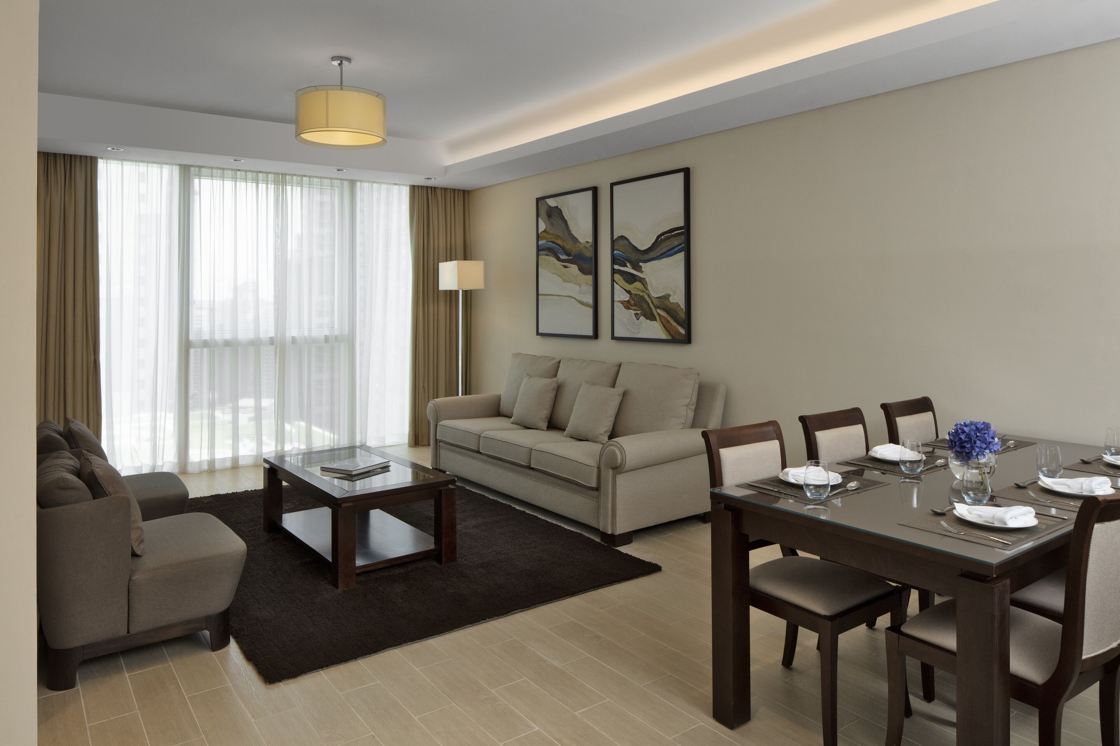 A modern hotel room with a sofa, a dining table set for four, a coffee table, and a large window with curtains.