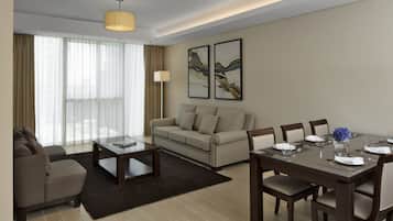 A modern hotel room with a sofa, a dining table set for four, a coffee table, and a large window with curtains.