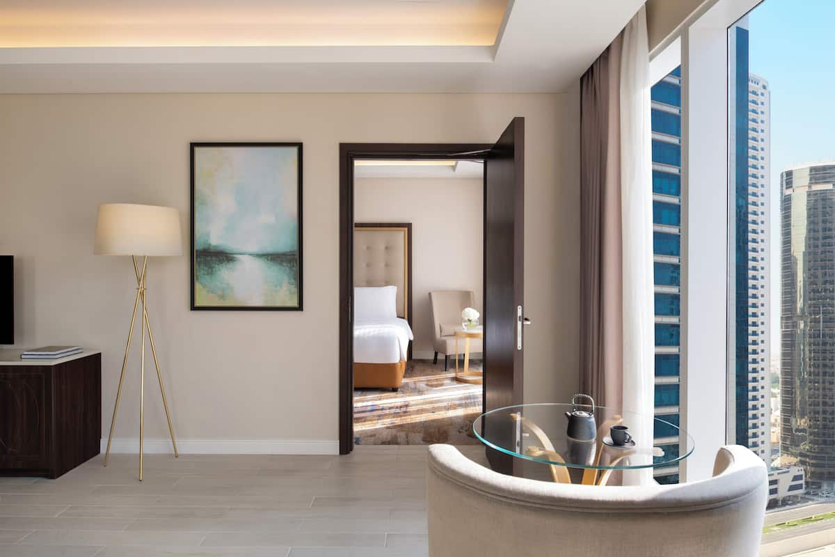 A modern hotel room with a large window, a glass table, a bed, and a view of the cityscape.