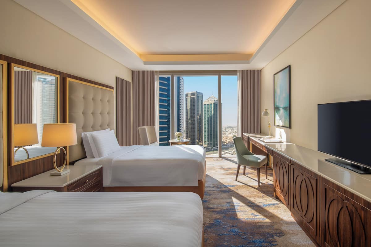 A hotel room with two beds, a desk, a TV, and a view of tall buildings.