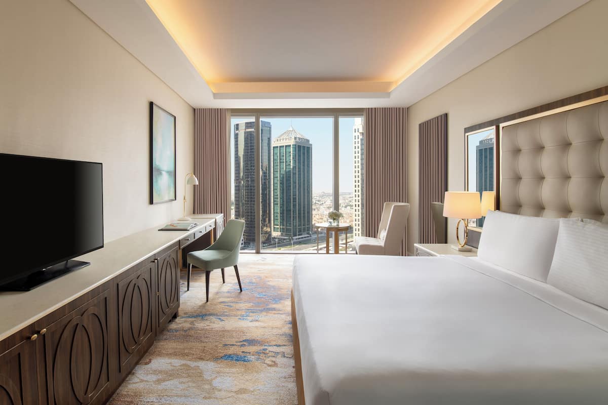 A hotel room with a large bed, a desk with a TV, a chair, and a view of tall buildings.