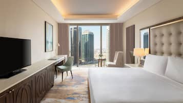 A hotel room with a large bed, a desk with a TV, a chair, and a view of tall buildings.