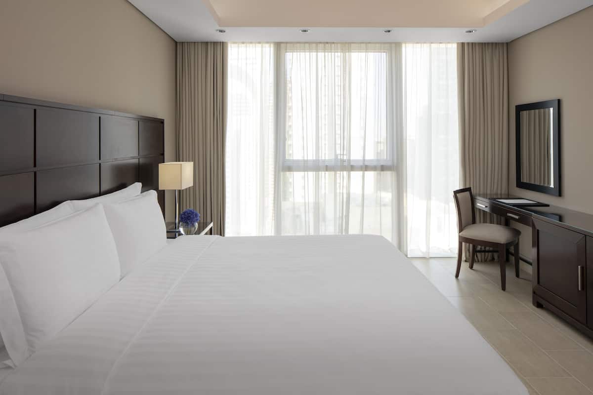 A hotel room with a large bed, a desk, a chair, a lamp, and a large window with curtains.