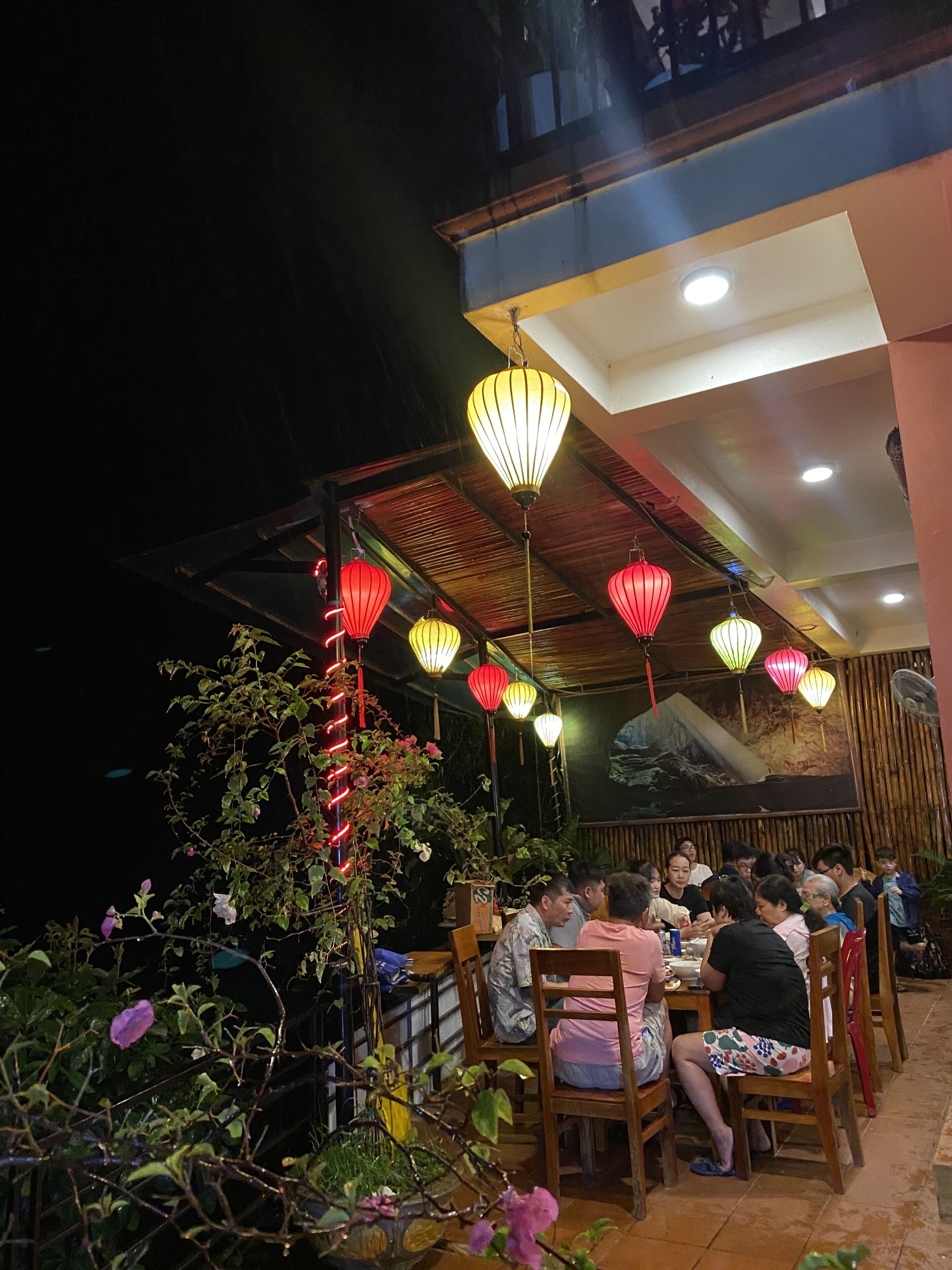 Un área de comedor al aire libre con mesas y sillas, rodeada de plantas y faroles decorativos.