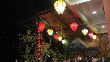 Un área de comedor al aire libre con mesas y sillas, rodeada de plantas y faroles decorativos.