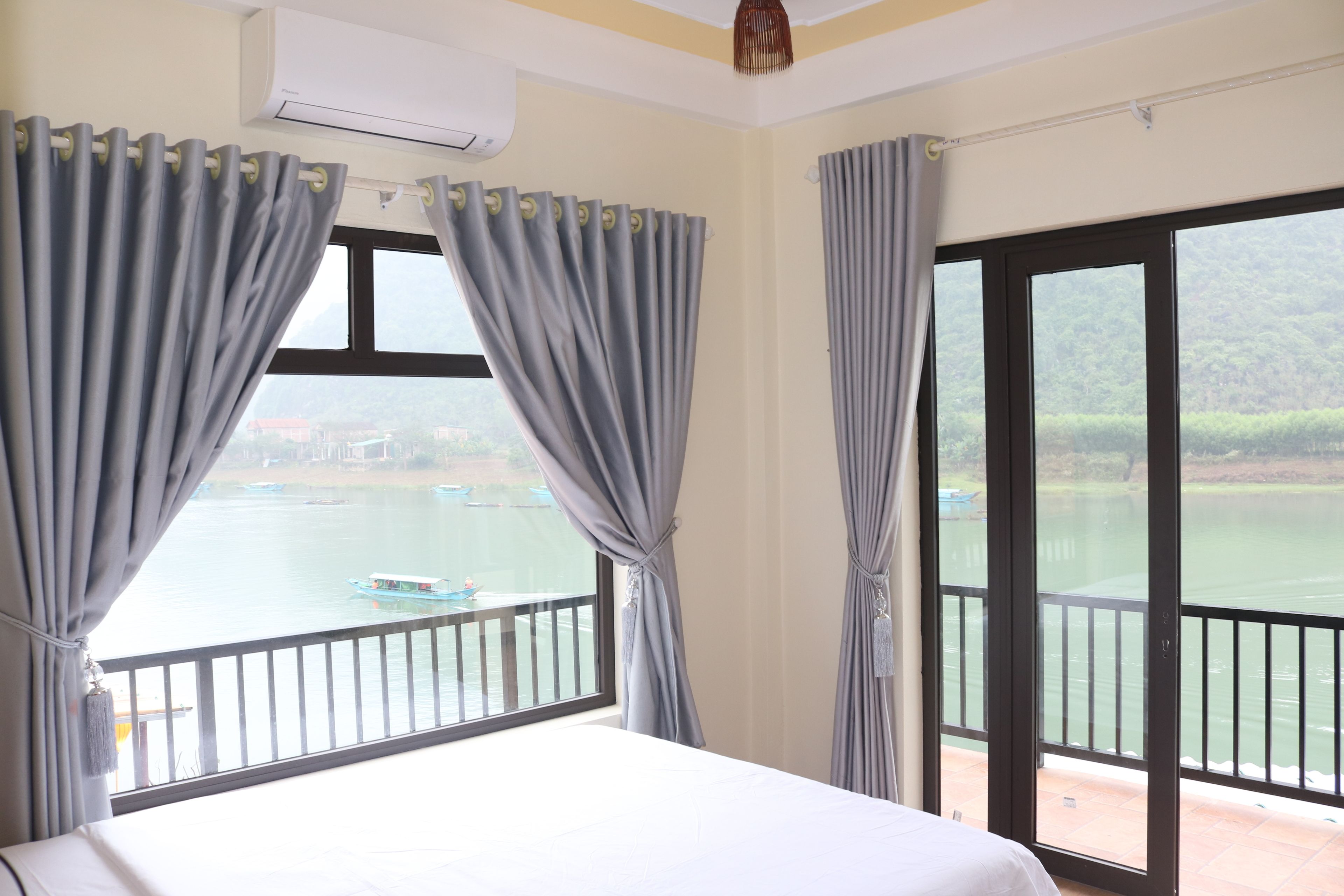 A hotel room with a bed, a window with curtains, an air conditioning unit, and a view of a body of water with a boat.