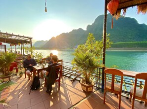 A terrace with a view of a lake and mountains, featuring tables and chairs, potted plants, and a thatched roof structure. - Phong Nha Coco Riverside Homestay (Bo Trach)